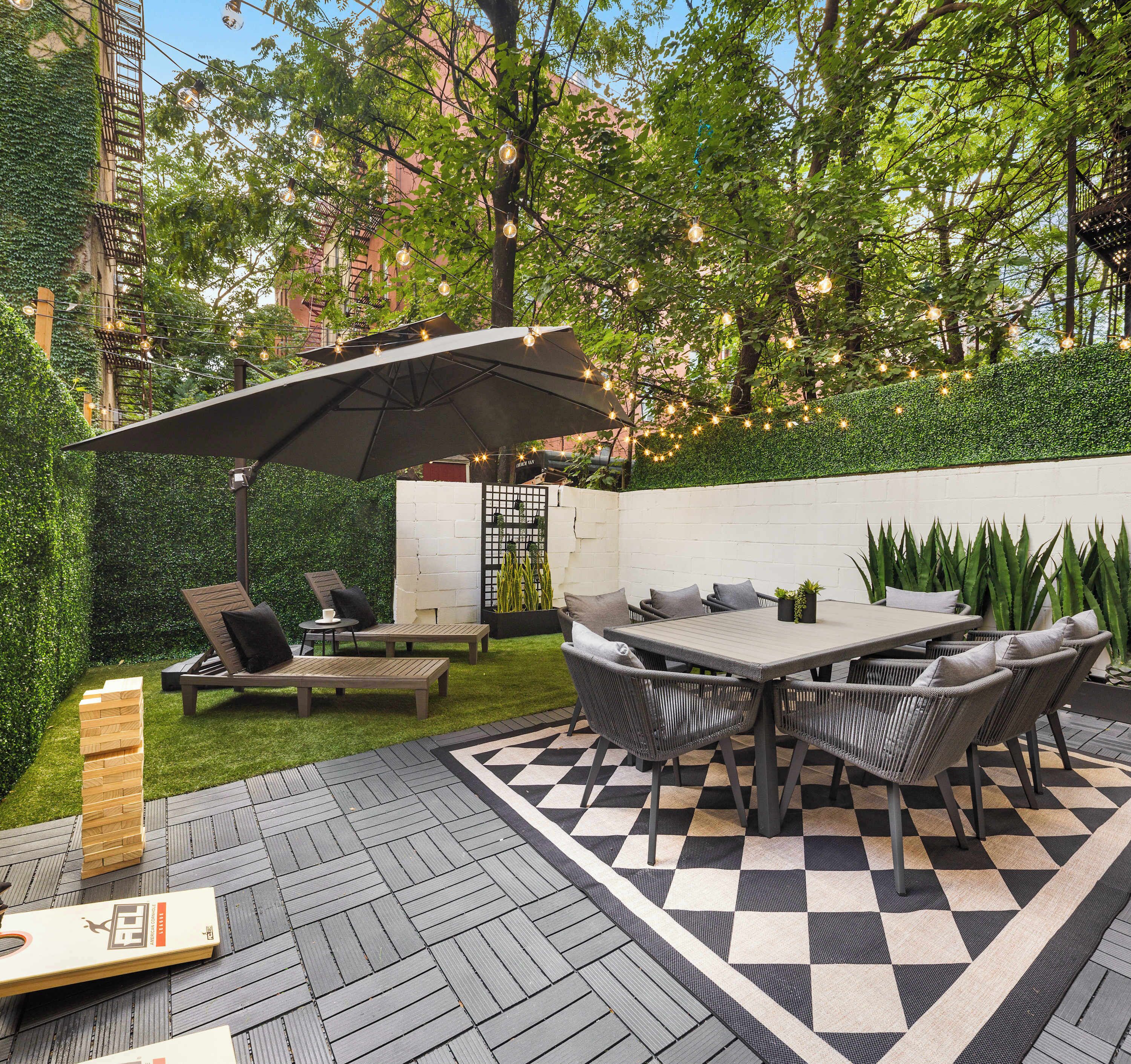 Un patio exterior con suelo a cuadros, una mesa con sillas y una pérgola con luces colgantes.