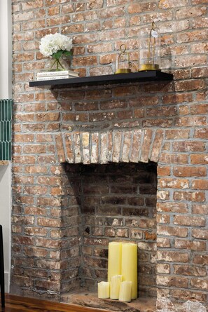 A brick fireplace with a shelf above it holding books and a vase with flowers. - Harlem Garden Retreat | Sleeps 10 & Private Patio! (New York)