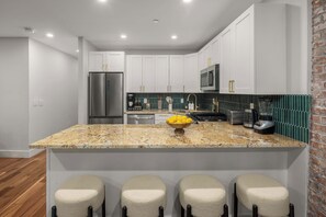 A modern kitchen with white cabinets, a granite countertop, a stainless steel refrigerator, a microwave, and a gas stove. There are three bar stools at the island.
