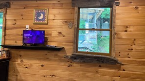 Interior de una cabaña de madera con un televisor en un estante, un cuadro en la pared y una ventana con vistas a una estructura de madera.