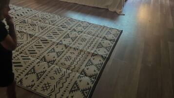 Um quarto com teto inclinado, uma cama grande com roupa de cama estampada, uma janela com vista para o verde e uma mesinha de cabeceira com uma luminária.