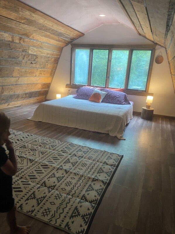 A bedroom with a sloped ceiling, a large bed with patterned bedding, a window with a view of greenery, and a small bedside table with a lamp.