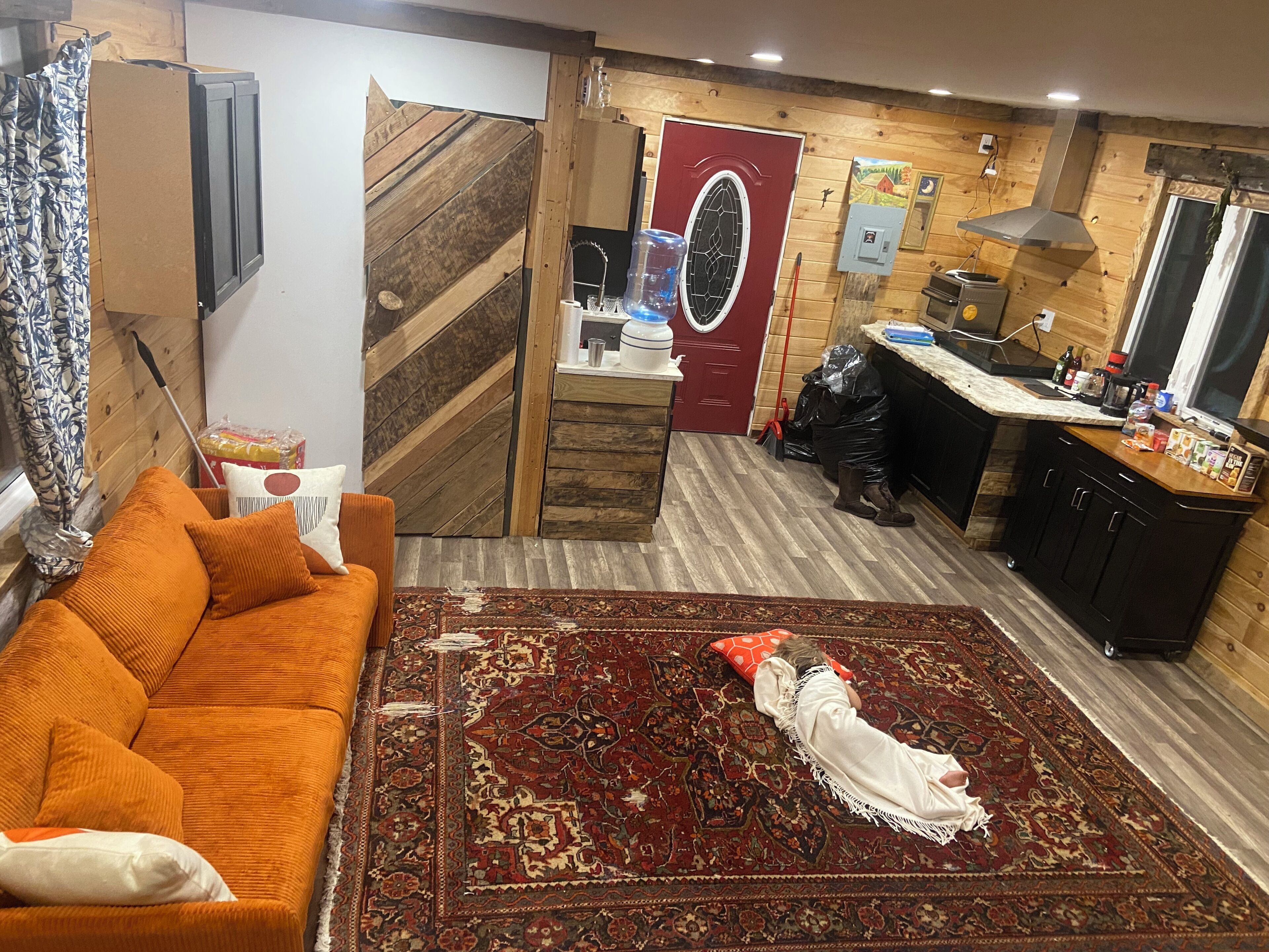 Sala de estar com sofá, tapete estampado, parede de madeira, área de cozinha com armários e uma porta vermelha.