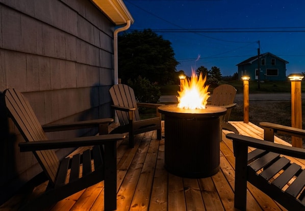 A wooden deck with a central fire pit, surrounded by chairs and a table.