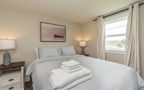 Una cama bien hecha con ropa de cama a rayas, un cuadro de paisaje enmarcado encima y una ventana con cortinas a la derecha.