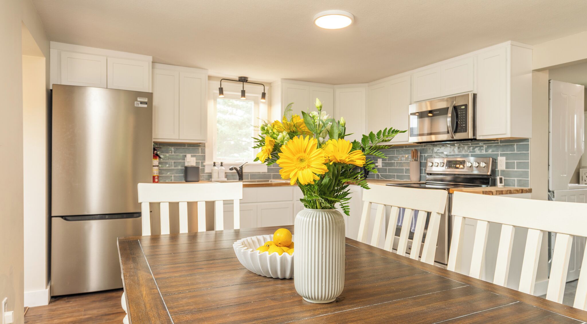 A modern kitchen with white cabinetry, stainless steel appliances, and a wooden dining table with a vase of yellow flowers.