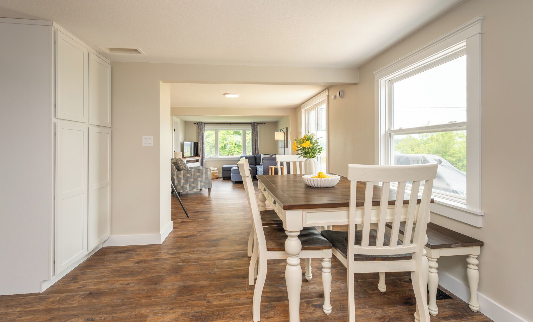 A spacious dining area with a long wooden table and white chairs, leading to a living room with a sofa and a television.