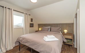 A bedroom with a bed, two bedside lamps, a window with curtains, a wooden headboard, and a wall with two framed pictures.