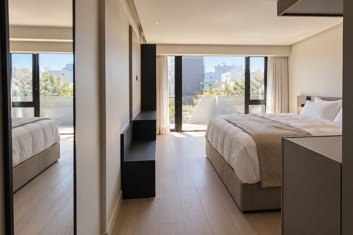 A modern bedroom with a large bed, a built-in desk, and a view of the city through large windows.