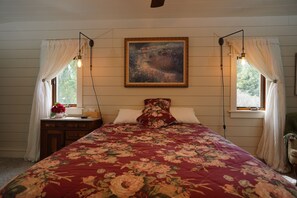 Ein Schlafzimmer mit einer blumigen Bettdecke, einem Bild über dem Bett und zwei mit Vorhängen versehenen Fenstern.