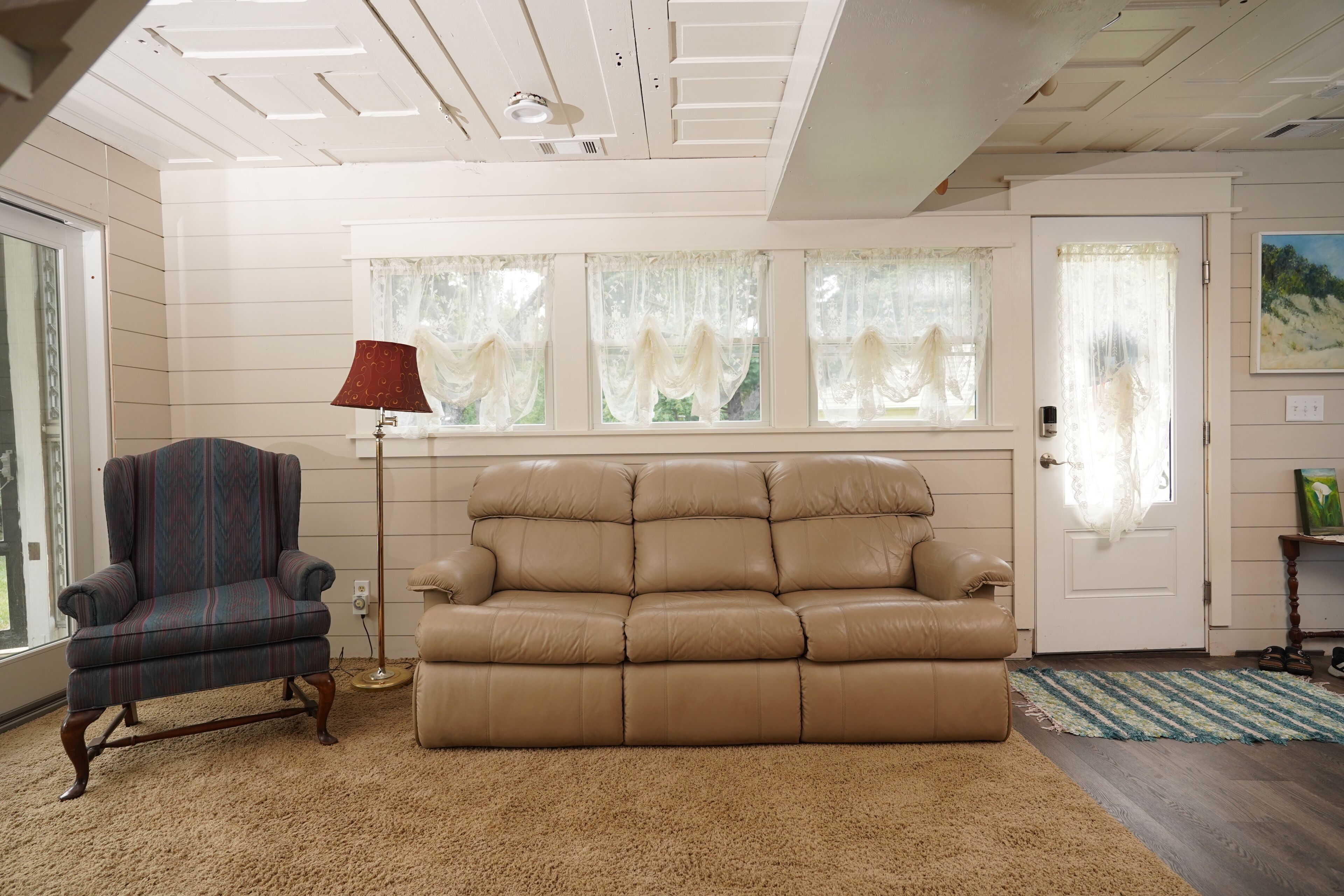 Una habitación con un sofá de cuero color beige, un sillón estampado, una lámpara de pie roja y una puerta con un cuadro encima.