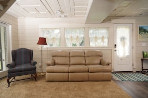 Ein Zimmer mit einem beigefarbenen Ledersofa, einem gemusterten Sessel, einer roten Stehlampe und einer Tür mit einem Bild darüber.