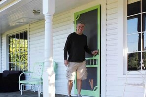 A person standing on a porch with a green door, white siding, and a chair.