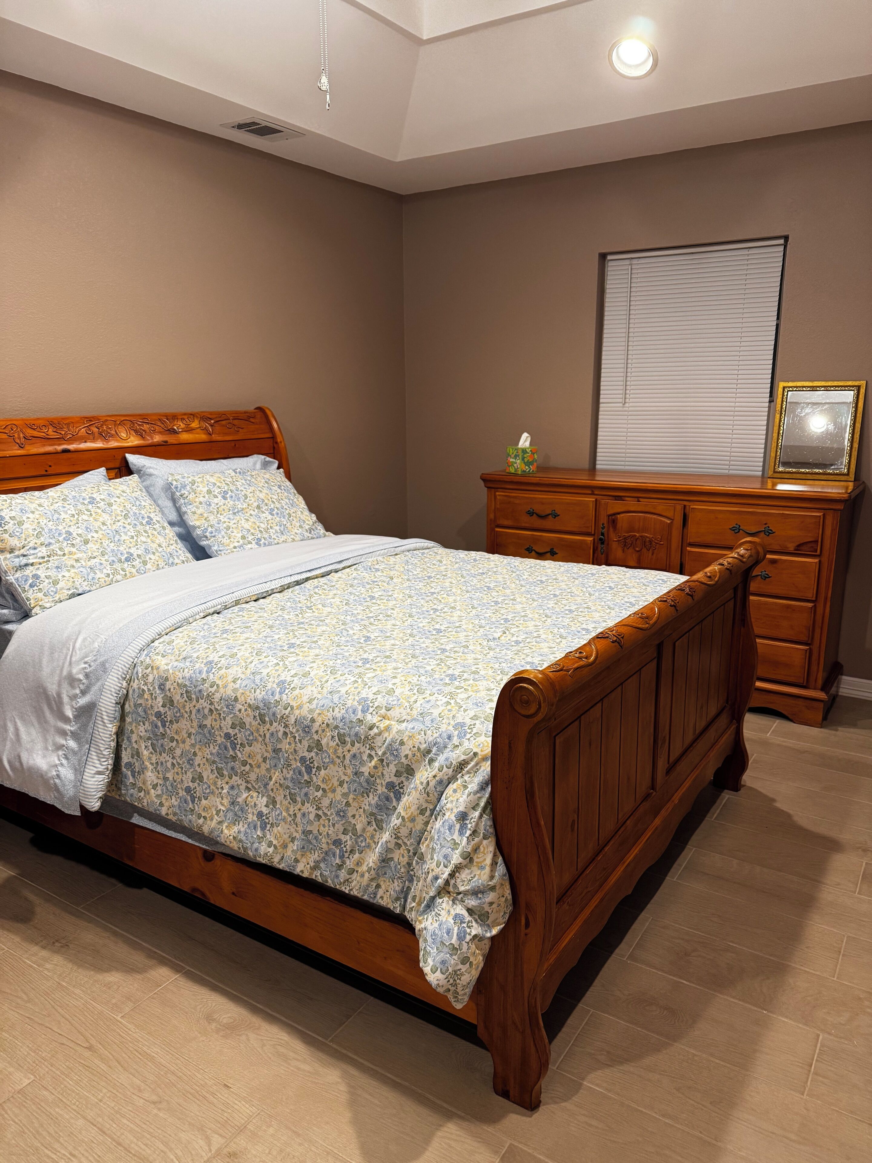 A bedroom with a wooden bed, a dresser, a mirror, and a window with blinds.