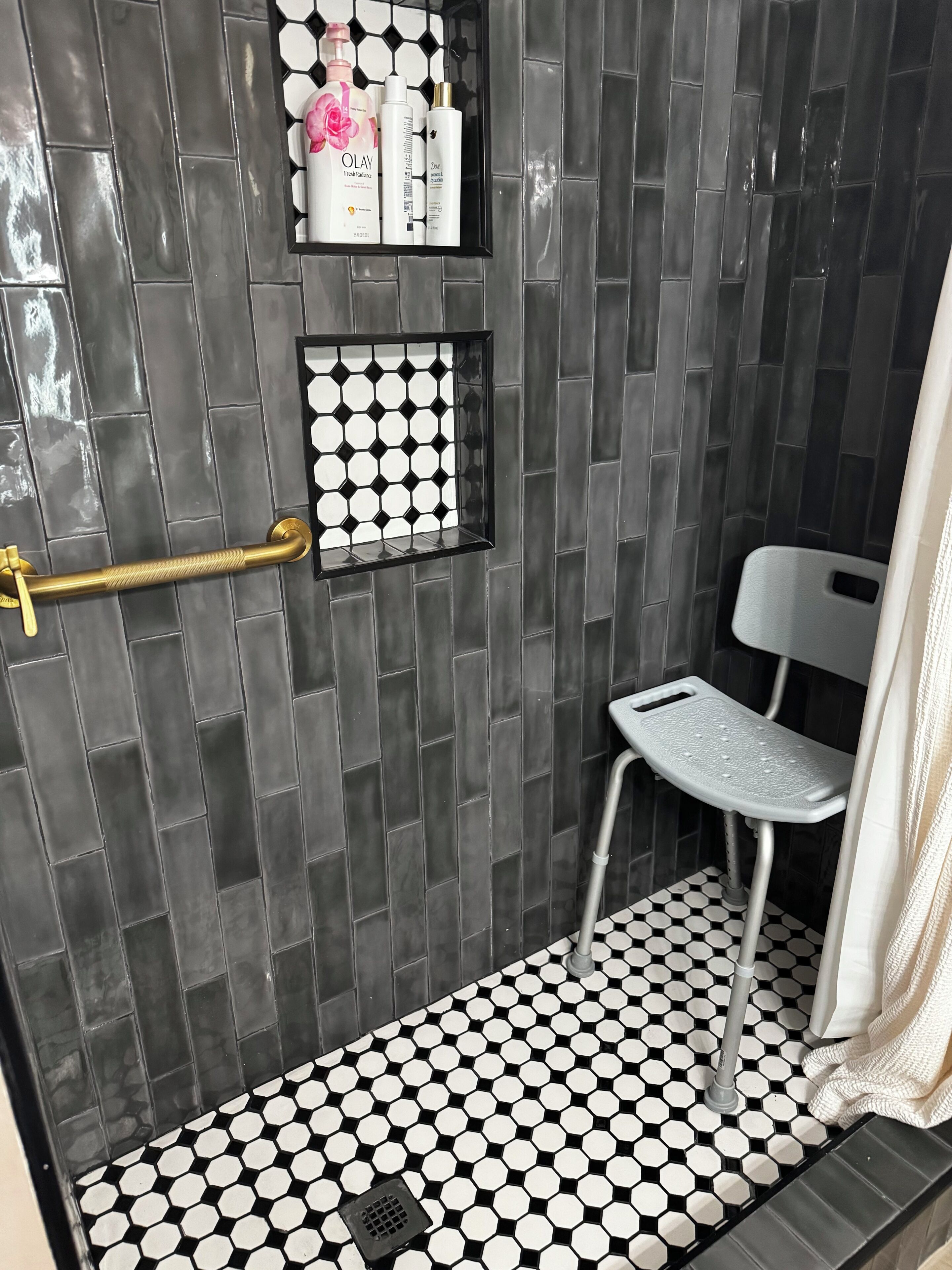 A bathroom with a black and white hexagonal tile floor, a shower with a black tiled wall, a white chair, and a shelf with toiletries.