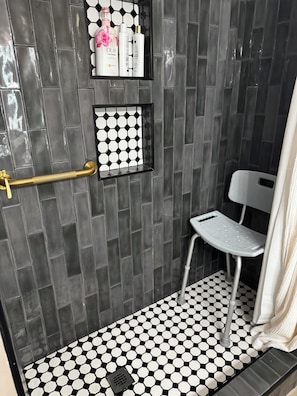 Une salle de bain avec un sol carrelé de motifs hexagonaux noirs et blancs, une douche avec un mur carrelé de carreaux noirs, une chaise blanche et une étagère contenant des articles de toilette.