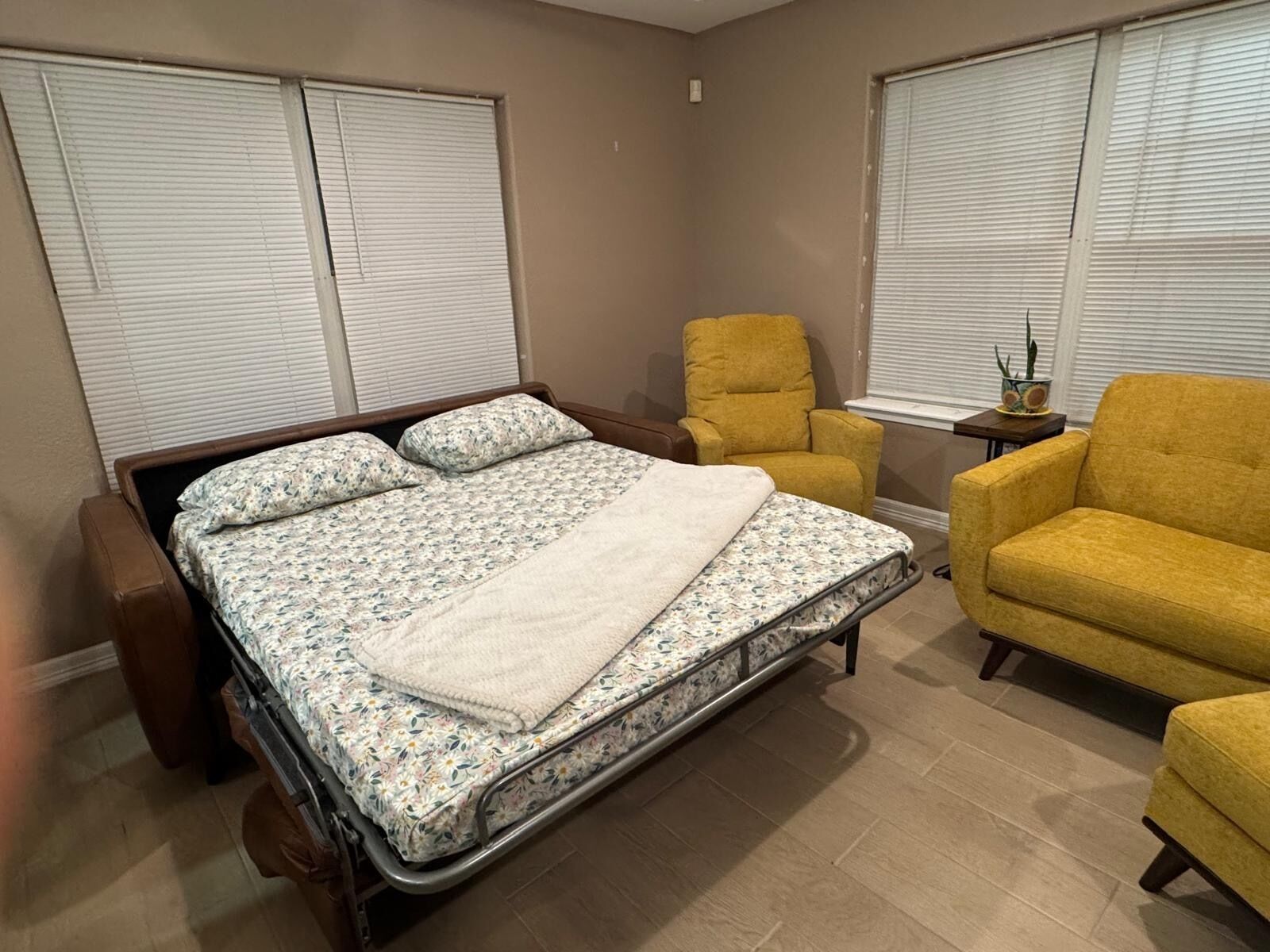 A bedroom with a bed, two chairs, a bedside table with a plant, and a window with blinds.