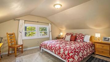 Um quarto com teto inclinado, uma cama com colcha florida, uma mesinha de cabeceira de madeira, uma cadeira e uma janela com vista para árvores.