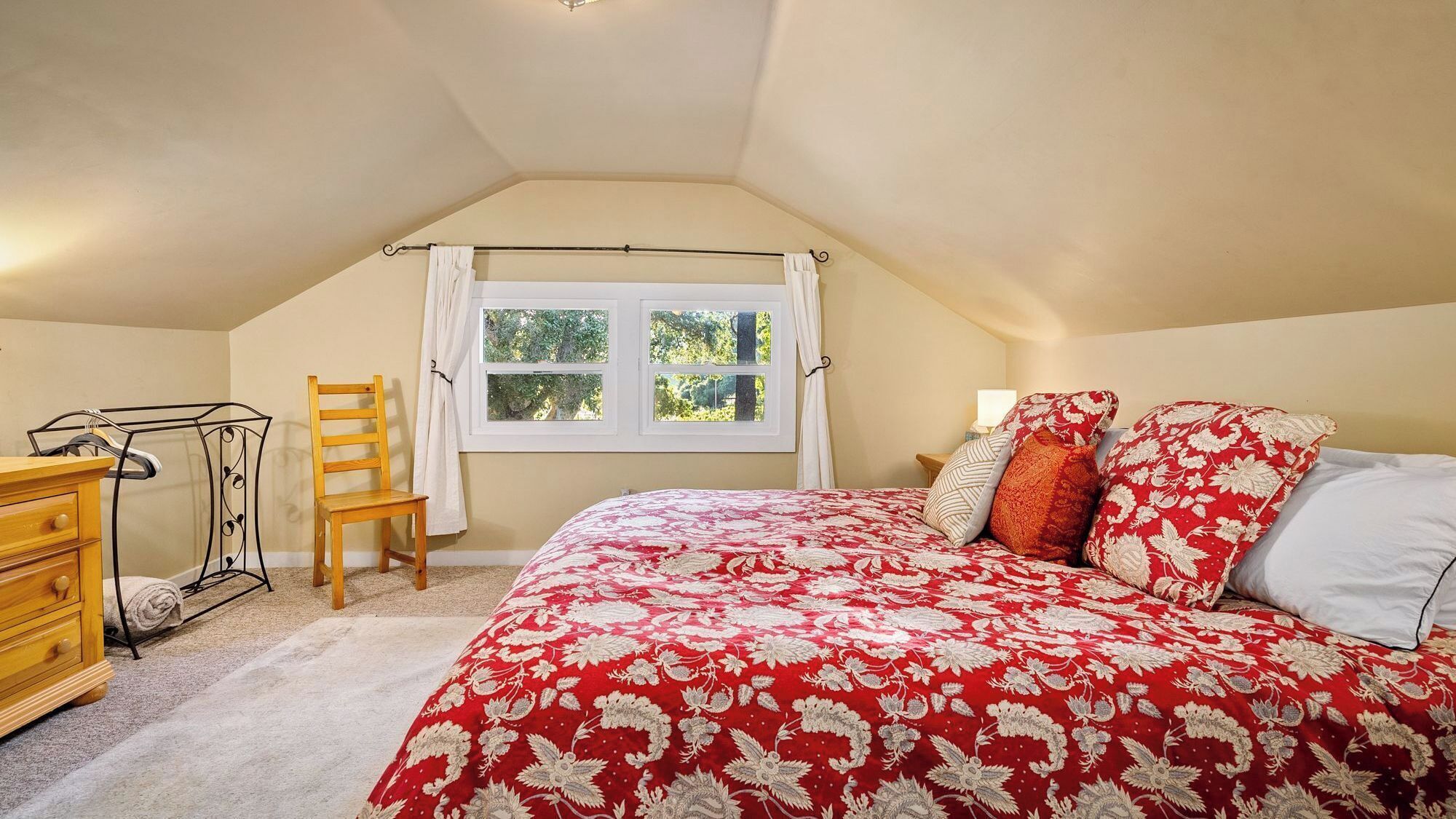 A bedroom with a sloped ceiling, a bed with a red floral bedspread, a wooden chair, a metal chair with a towel, and a window with curtains.