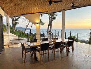 A dining area with a view of the sea, featuring a wooden table and chairs, a ceiling fan, and a staircase leading to an outdoor terrace. - Seataya Palace - 3-BDR Ocean View Luxury Villa & Infinity Pool (Santa Teresa)