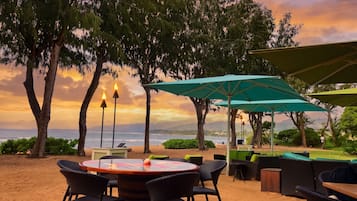 A beachside outdoor seating area with a round table and chairs, surrounded by trees and overlooking the sea.