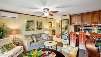 A living room with a sofa, coffee table, and a ceiling fan. There is a kitchen area with wooden cabinets and a dining table.