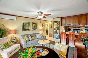 A living room with a sofa, coffee table, and a ceiling fan. There is a kitchen area with wooden cabinets and a dining table. - Kapaa Kauai 1BR Condo: Walk to Beach, Pool & A/C. (Kapaʻa)