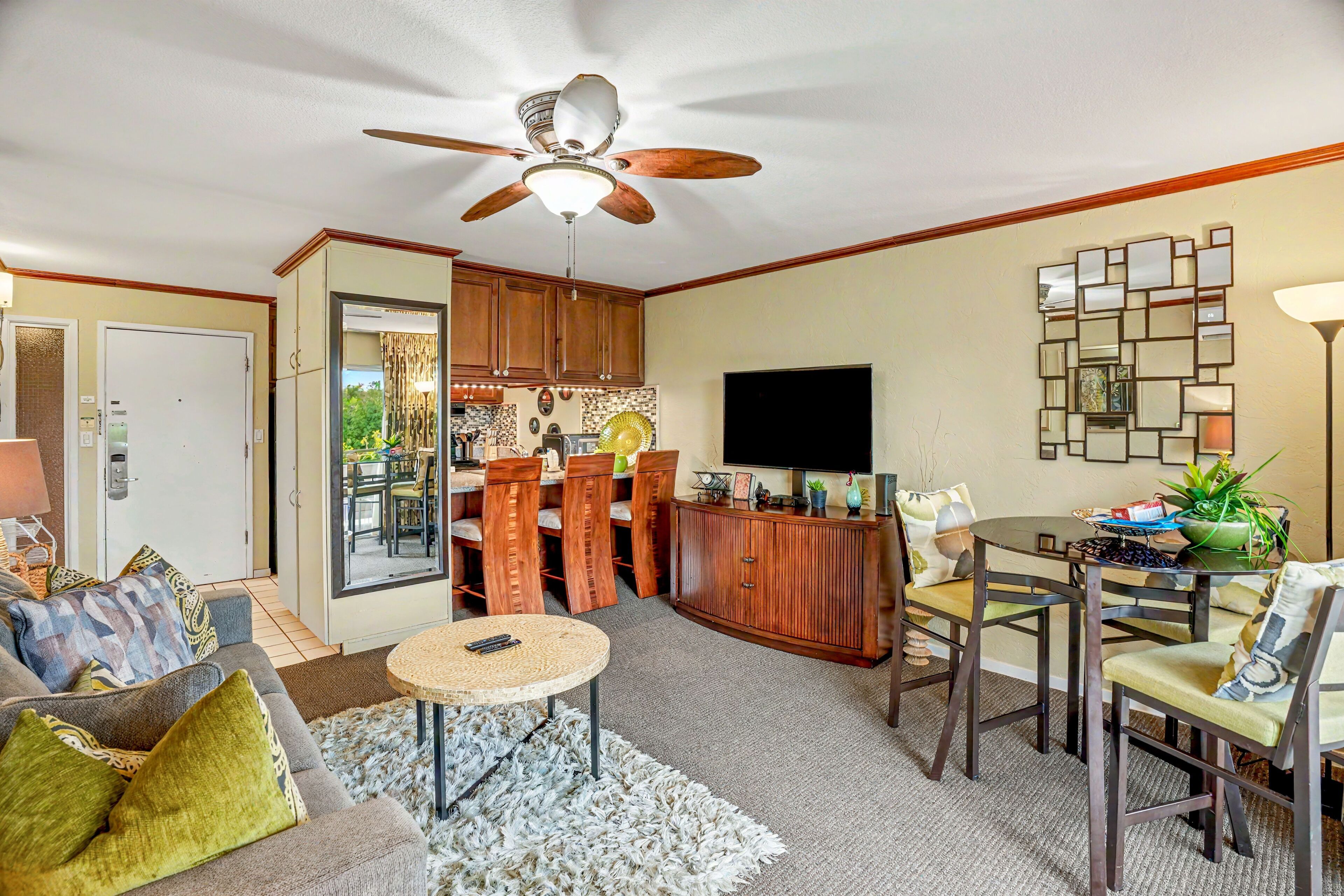 A living room with a ceiling fan, a TV, a dining area with chairs, a kitchen with cabinets, and a door leading to another room.