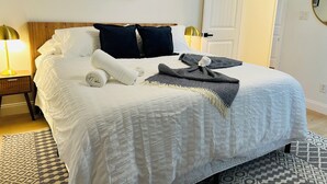 Ein ordentlich bezogenes Bett mit einer weißen Decke, zwei dunklen Kissen und einem gefalteten weißen Handtuch. Auf dem Bett liegt eine Decke mit Muster.