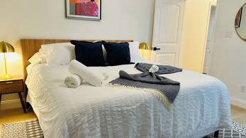 A neatly made bed with a white comforter, two dark pillows, and a folded white towel. There is a patterned throw blanket on the bed.