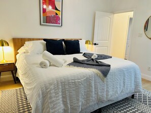 Una cama bien hecha con edredón blanco, dos almohadas oscuras y una toalla blanca doblada. Hay una manta decorativa estampada sobre la cama.