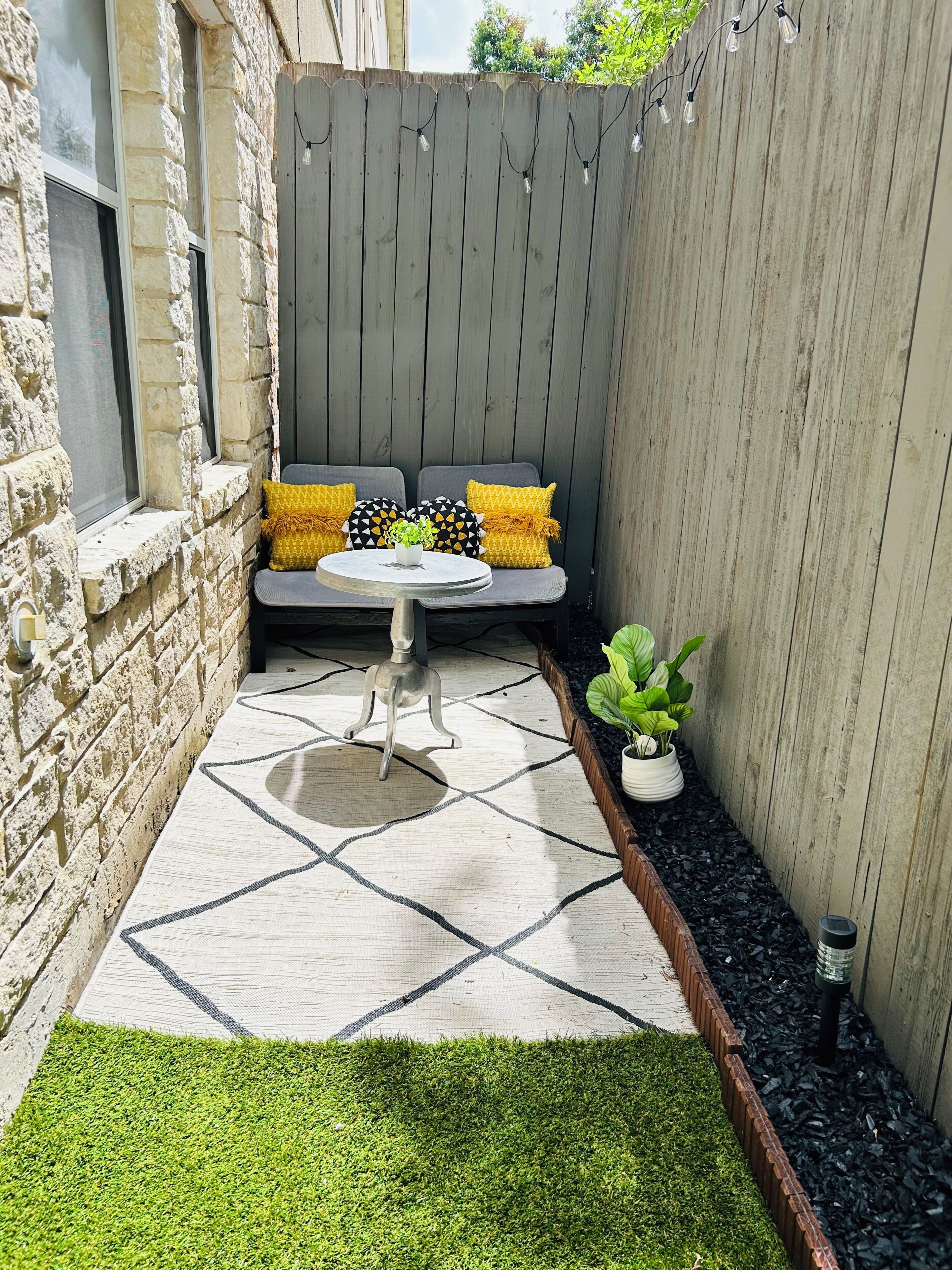 A patio area with a stone wall, a wooden fence, a seating bench with cushions, a small table, and a potted plant.