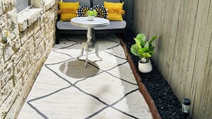 Eine Terrasse mit Steinmauer, Holzlattenzaun, Sitzbank mit Kissen, einem kleinen Tisch und einem Blumentopf.