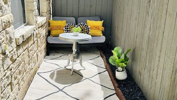 A patio area with a stone wall, a wooden fence, a seating bench with cushions, a small table, and a potted plant.