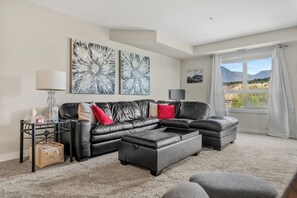 A modern living room with a black leather sofa, a coffee table, and a view of mountains through a large window. - Daybreak | Outdoor Pool | Hot Tub | Gym | Balcony (Invermere)