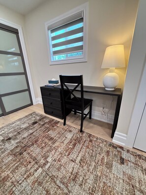 Een kamer met een bureau, stoel en lamp. Er is een raam met jaloezieën, een vloerkleed en een deur.