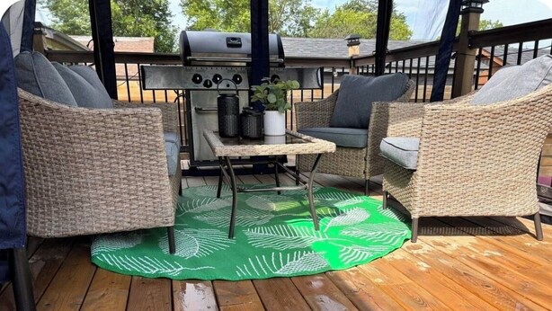 Una terraza techada al aire libre con mobiliario de mimbre, una parrilla y un tapete verde con estampado.