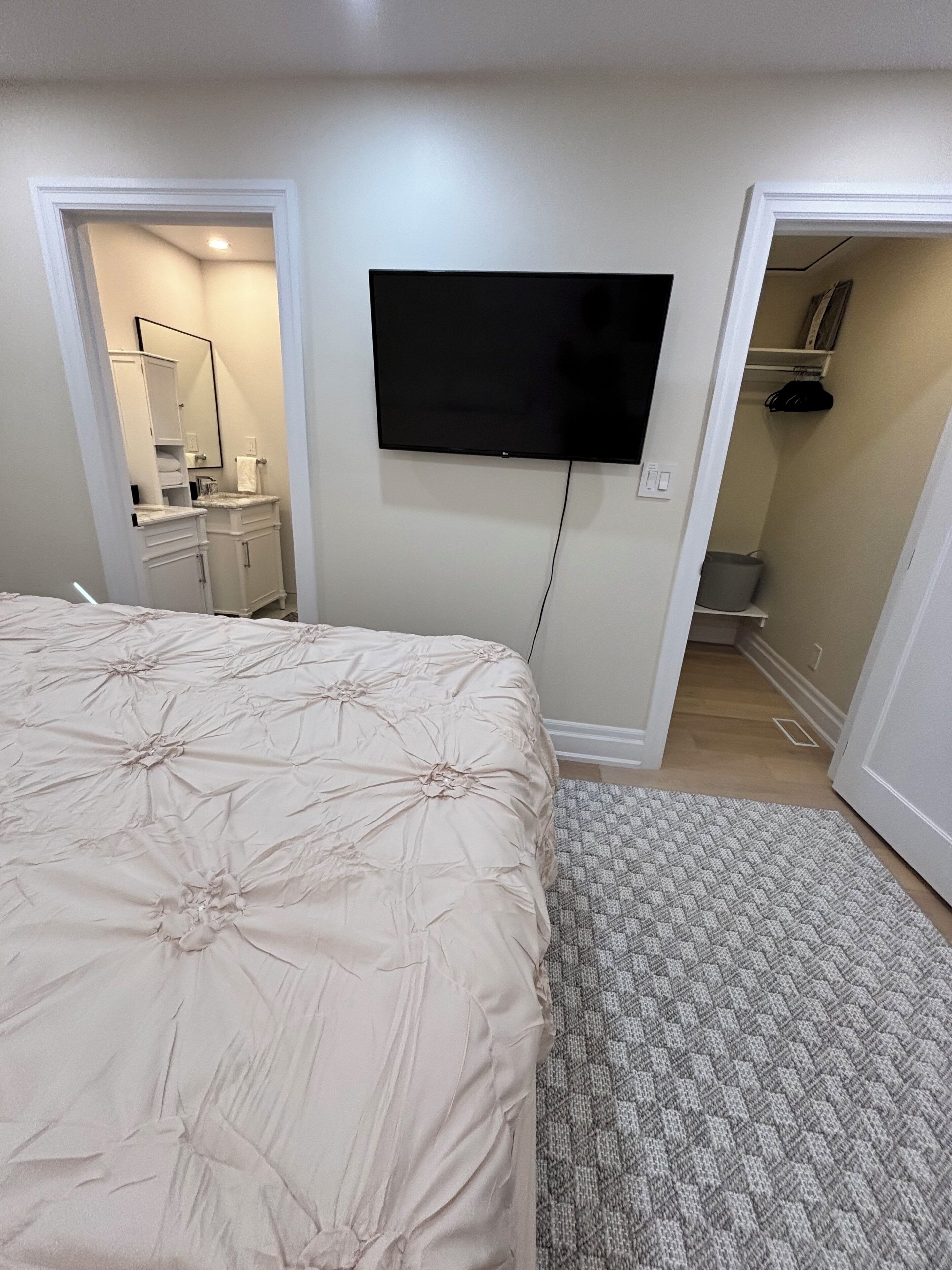 A bedroom with a bed, a television mounted on the wall, a bathroom with a mirror, and a hallway leading to another room.