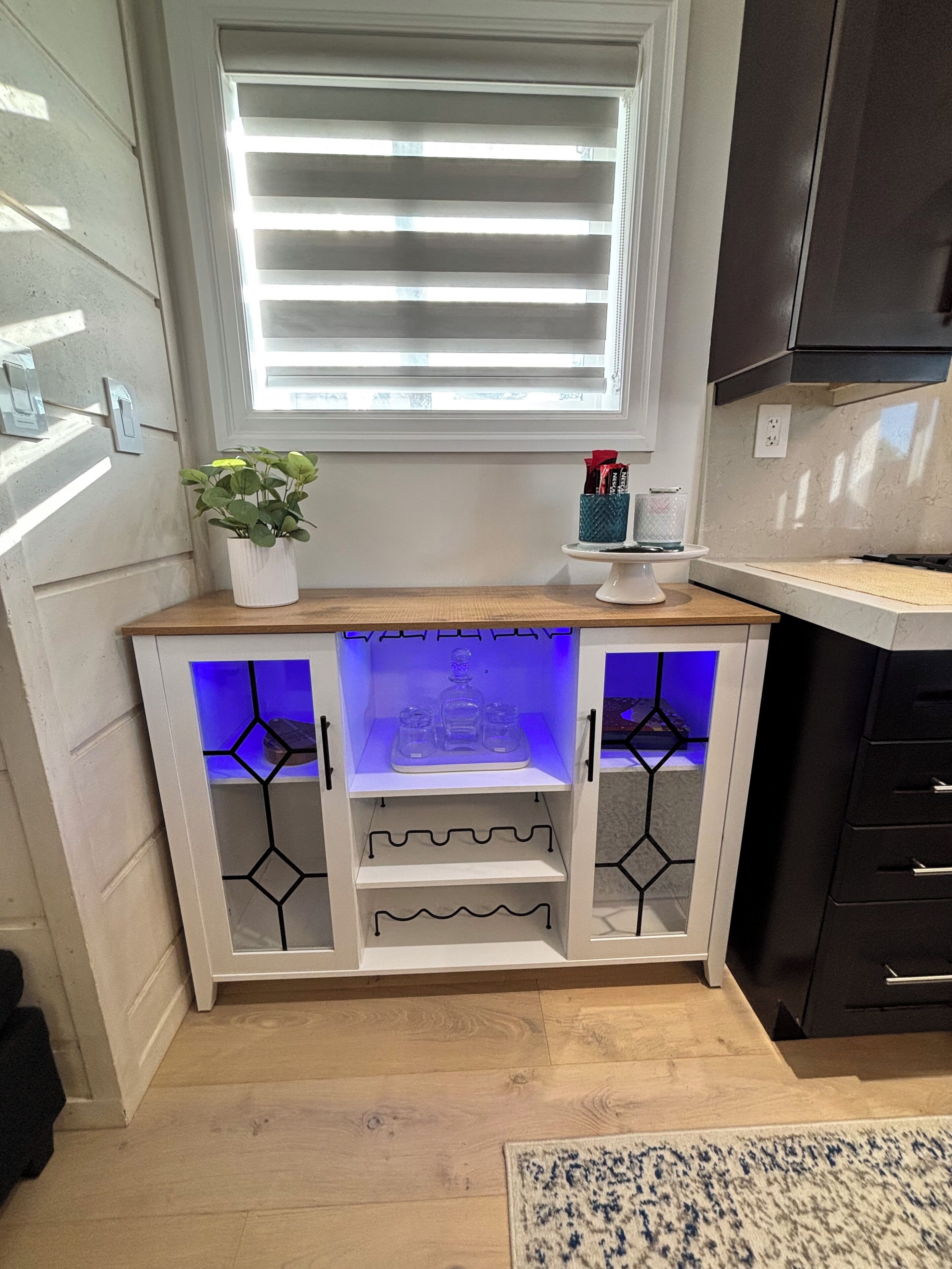Una cocina con un armario blanco empotrado, una ventana con persianas, una planta en maceta y un estante pequeño con objetos.