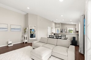 A modern living room with a sectional sofa, a dining area with a table and chairs, and a kitchen area with a refrigerator and microwave.