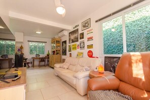 A living room with a sofa, a coffee table, and a dining area with a table and chairs. There are wall decorations and a window with a view of greenery.