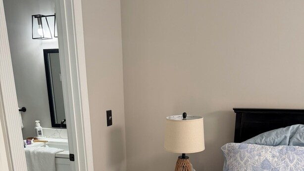 Une chambre à coucher avec un lit, une table de chevet avec une lampe, une petite armoire et une salle de bain visible à travers une porte ouverte.