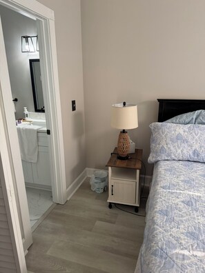 A bedroom with a bed, a nightstand with a lamp, a small cabinet, and a bathroom visible through an open door. - Myrtlewood Golf Getaway - Renovated 2BR Condo Near Beach (Myrtle Beach)
