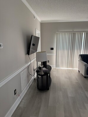 A modern living room with a flat-screen TV mounted on the wall, a lamp on a side table, and a large window with vertical blinds. - Myrtlewood Golf Getaway - Renovated 2BR Condo Near Beach (Myrtle Beach)