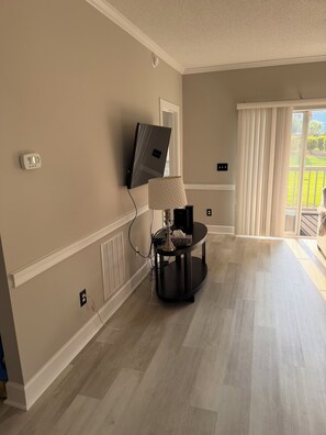 A room with a flat-screen TV mounted on the wall, a lamp on a small table, and a sliding glass door leading to an outdoor area. - Myrtlewood Golf Getaway - Renovated 2BR Condo Near Beach (Myrtle Beach)