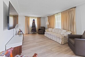 A living room with a flat-screen TV, a sofa, a chair, a small table with a lamp, and a Christmas tree. - Trinity Walk 4 (Steubenville)