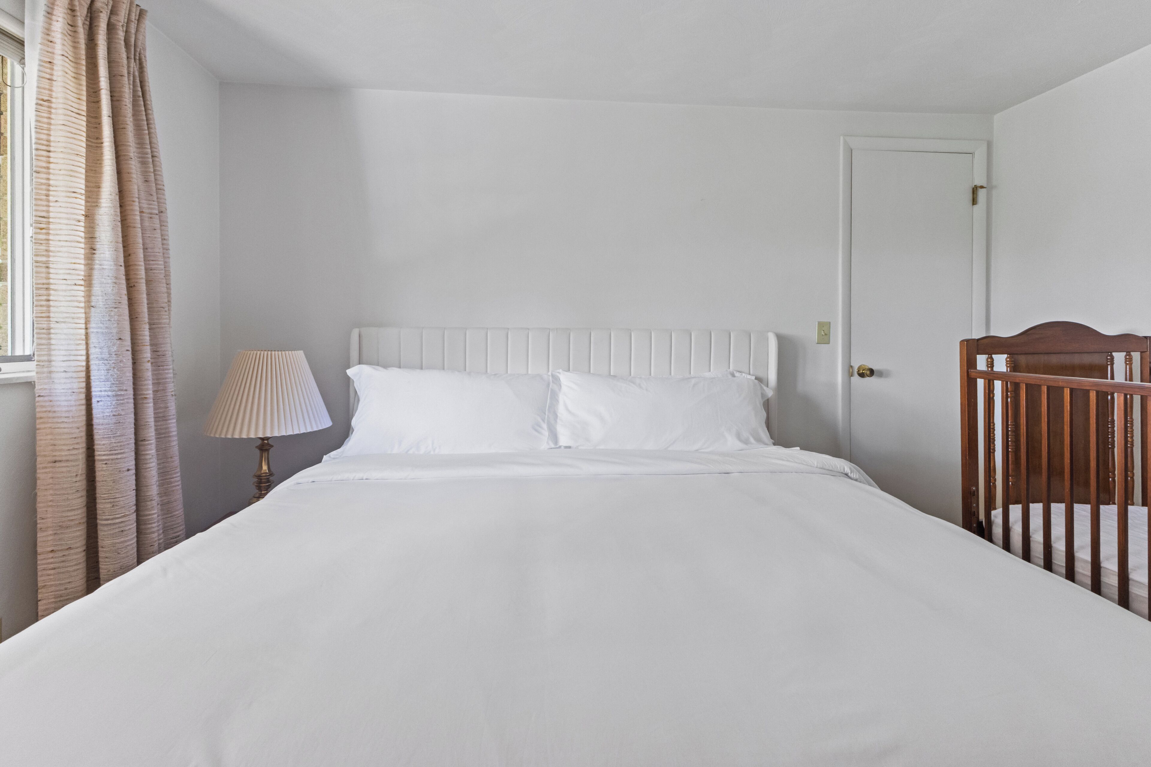 A single bed with white bedding, a bedside lamp, and a wooden crib in a room with white walls and a door.