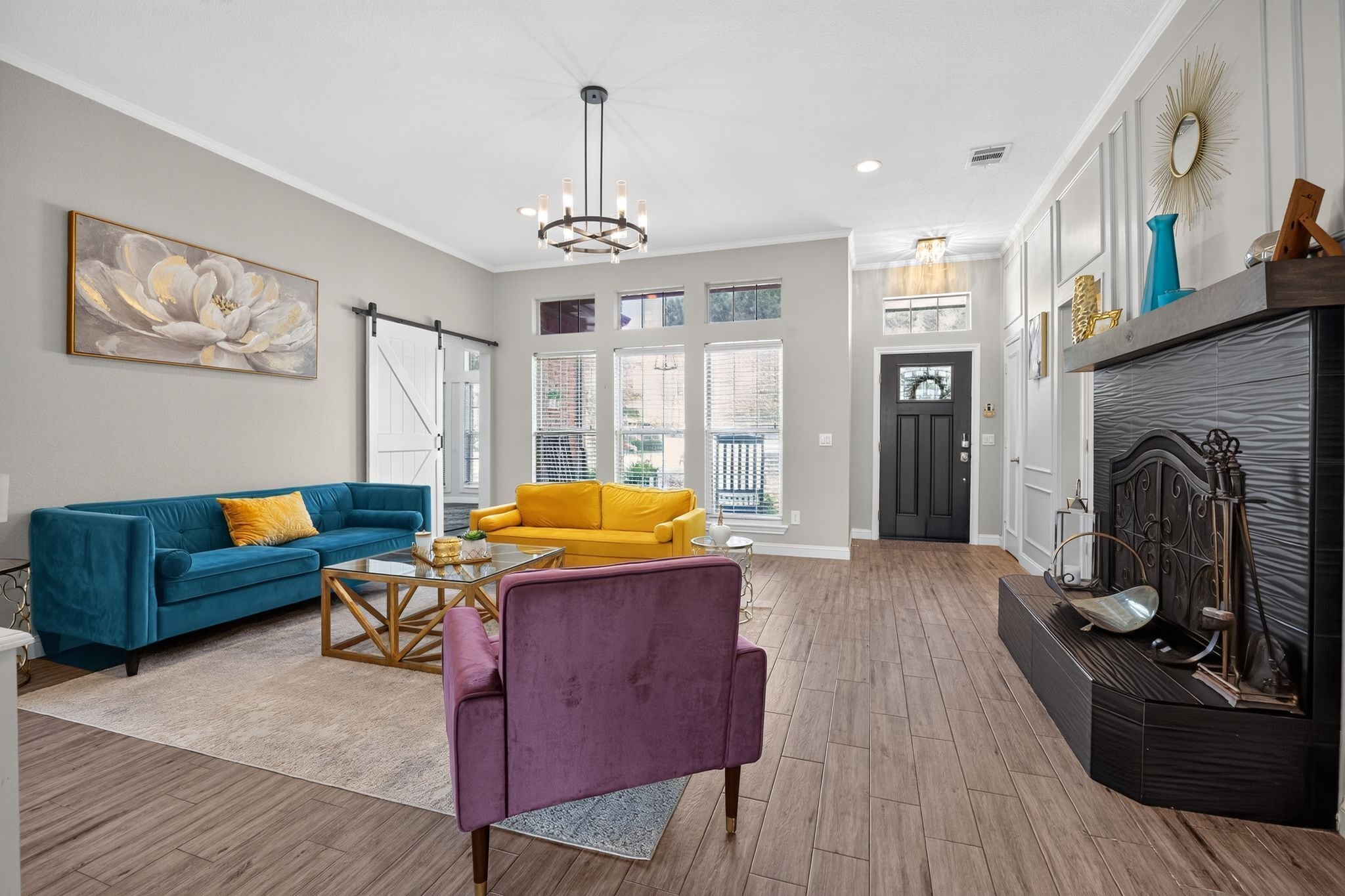 A spacious living room with a blue sofa, a yellow armchair, and a purple chair. There is a fireplace with decorative items and a large window with blinds.