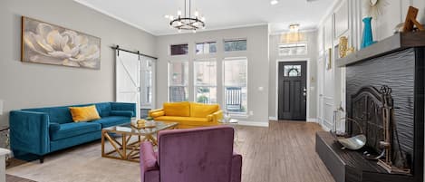 A spacious living room with a blue sofa, a yellow armchair, and a purple chair. There is a fireplace with decorative items and a large window with blinds.