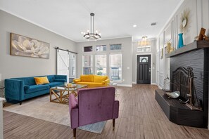 Ein großzügiger Wohnraum mit einem blauen Sofa, einem gelben Sessel und einem violetten Stuhl. Ein Kamin mit Dekorationen und ein großes Fenster mit Jalousien sind ebenfalls vorhanden.